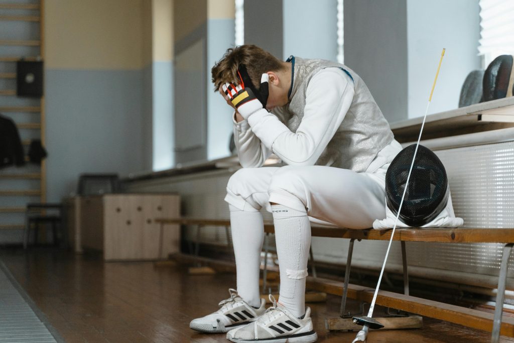 Young fencer with head in hands, showing frustration during indoor training session.